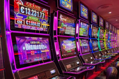 Máquinas caça-níqueis coloridas alinhadas em um cassino iluminado, com painéis de jackpot ao fundo.
