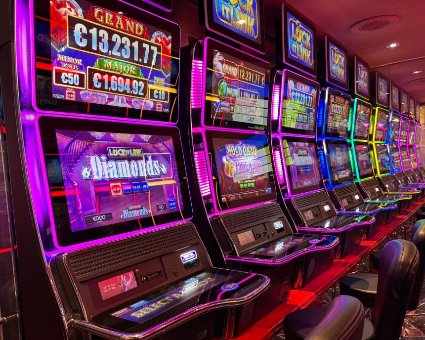 Máquinas caça-níqueis coloridas alinhadas em um cassino iluminado, com painéis de jackpot ao fundo.