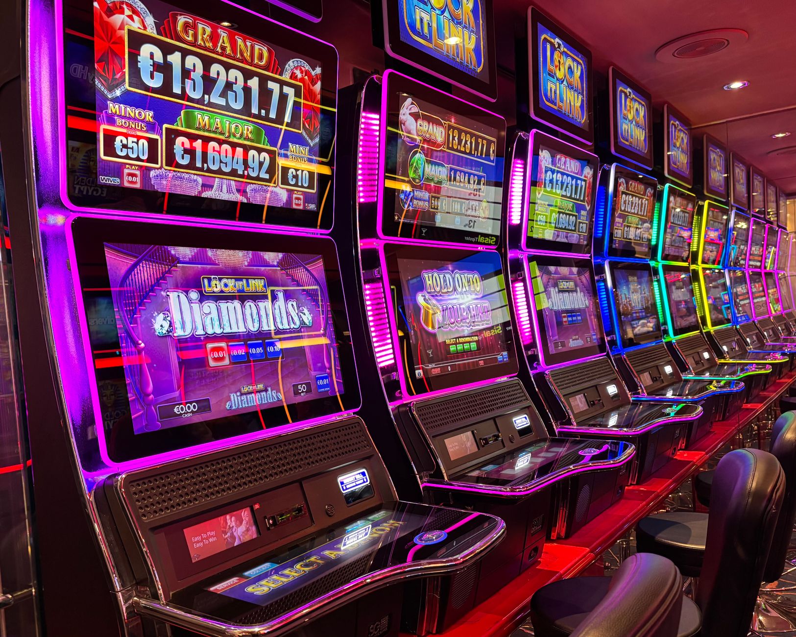 Máquinas caça-níqueis coloridas alinhadas em um cassino iluminado, com painéis de jackpot ao fundo.