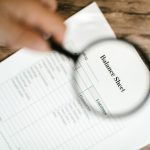 Balanço financeiro examinado com uma lupa sobre uma mesa de madeira.