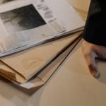 Mão apoiada sobre uma mesa ao lado de documentos de investigação e recortes de jornal.