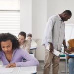 Estudantes de diferentes perfis fazem uma prova em uma sala de aula, sob supervisão de um professor.