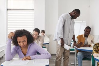 Estudantes de diferentes perfis fazem uma prova em uma sala de aula, sob supervisão de um professor.